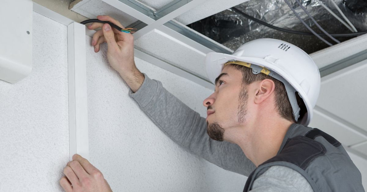 Electrician wearing a hard hat working on the bonding of cable trunking, connecting electrical wires to a ceiling-mounted conduit system.